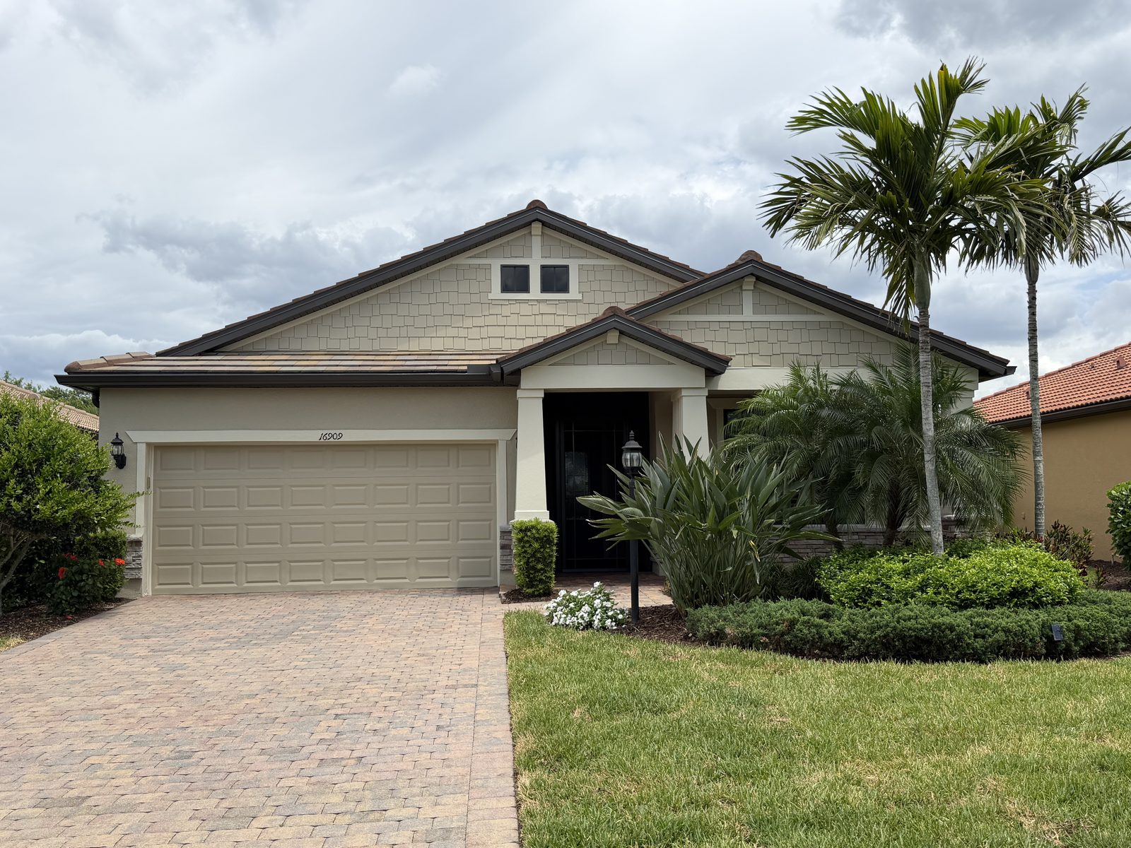 Single-story tan Florida home with a tile roof, decorative gable, two-car garage, and tall palms